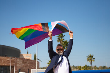 Mature gay man, executive, grey-haired, bearded, sunglasses, jacket and tie, waving the new lgbtiq+...