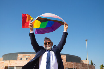 Mature gay man, executive, grey-haired, bearded, sunglasses, jacket and tie, waving the new lgbtiq+...