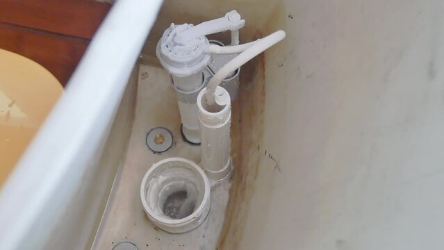 Leaking toilet tank with missing float ball and flapper mechanism failure. Toilet repair, home maintenance, plumbing accessories. View inside white ceramic toilet cistern, bowl, and intake valve.