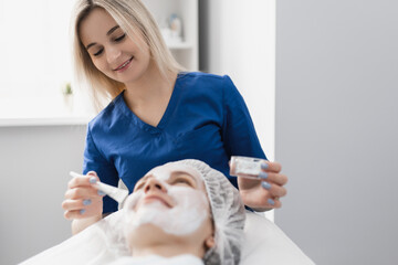 beautiful female cosmetologist applying a moisturizing white cream mask to her face with a brush to a smiling patient