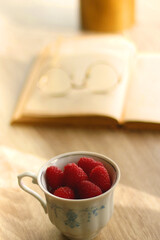 Porcelain cup filled with raspberries, open book, reading glasses and vase with flowers on the table. Selective focus.