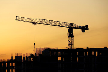 Silhouette of tower crane with cargo and workers on building scaffolding at sunset. Housing construction, apartment block in city on dramatic sky background