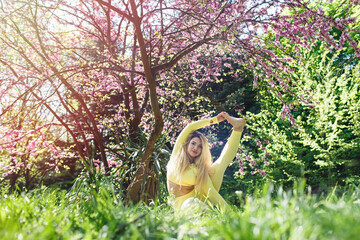 Young blonde woman in yellow sportswear does yoga or Pilates outdoors in nature. Attractive girl is sitting on a mat on grass in the park. Beautiful flowering pink trees in spring. Health and beauty.