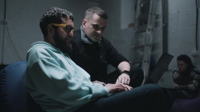 Handheld shot of a programmer with laptop and IT director of a company talking and discussing work together in a bomb shelter during the war