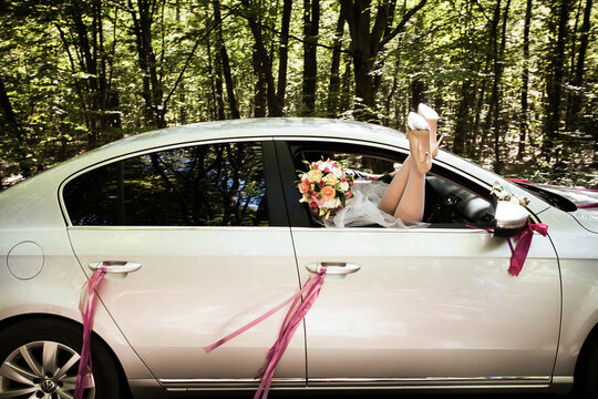 The bride put her bare legs out of the open car window, with a bouquet of roses in her hands. A car decorated with ribbons rides in the forest along the highway during the day.