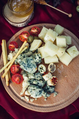 Composition of healthy food on a wooden tray, on a table with red material. Several varieties of cheese, including Roquefort, red tomatoes, pastries in the form of straws, honey, green olives.