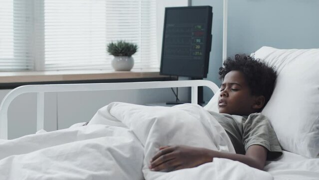 Side view of sick Black boy lying in hospital bed at daytime, breathing heavily
