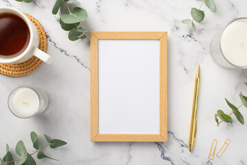 Obraz na płótnie Canvas Top view photo of wooden photo frame candles cup of tea on rattan serving mat gold pen clips and eucalyptus on white marble background with empty space