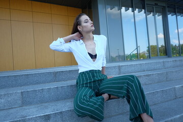 A young brunette girl in white shirt and striped trouser is sitting on the stairs