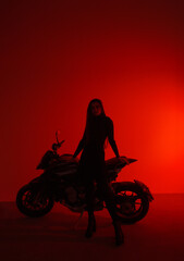 Naklejka na ściany i meble young teen brunette girl biker with long legs stands fashion like silhouette near black sports motorcycle on a red studio wall background. teen leisure. lifestyle fashion concept, free space