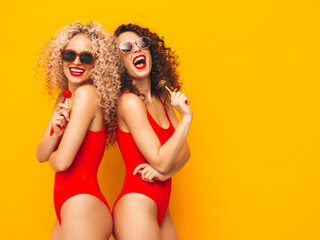 Two beautiful smiling hipster women in red swimwear bathing suits.Trendy models with curls hairstyle in studio. Hot female posing near yellow wall in sunglasses.Holding lollipop. Isolated on yellow - obrazy, fototapety, plakaty