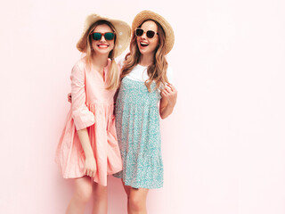 Two young beautiful smiling brunette hipster female in trendy summer dresses. Sexy carefree women posing near pink wall. Positive models having fun. Cheerful and happy. In hats