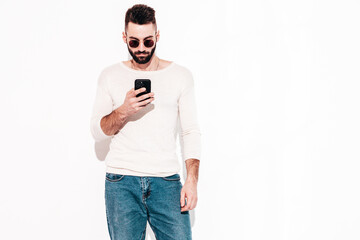 Handsome confident model.Sexy stylish man dressed in jacket and jeans. Fashion hipster male posing near white wall in studio. Holding smartphone. Looking at cellphone screen. Using apps