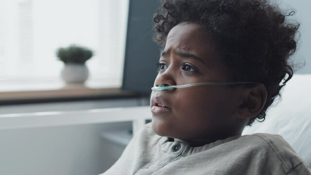 Close-up of African American boy with oxygen tube in nose sleeping in hospital bed at daytime, then waking up scared and calling mom