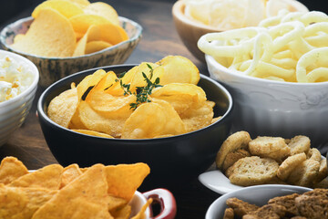 Unhealthy food or snacks. All classic potato snacks with peanuts, popcorn and onion rings and salted pretzels in bowl plates on old wooden background. Unhealthy food for figure, heart, skin, teeth.