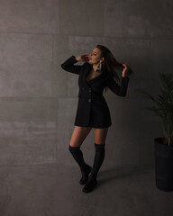 teen leisure. young girl in black slim dress-jacket and over the knee boots stands fashion and smiles with hands near hair on the gray loft wall background with plant . lifestyle concept, free space