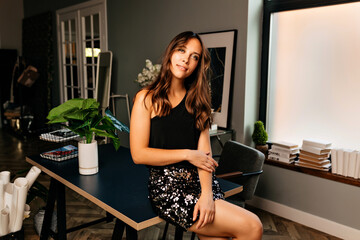 Portrait of stylish young woman in black elegant dress with bare shoulders. Pleased female caucasian model with dark curly hairstyle is sitting on the table at home