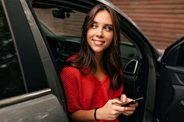 Obraz na płótnie Canvas Close up outdoor portrait of lovely cute girl with long wavy hair with wonderful smile is scrolling smartphone while sitting in the car