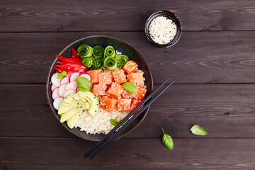 Poke bowl with salmon, rice, avocado, cucumber, radish, pepper, sesame seeds on dark wooden background. Close up. Hawaiian diet food with fish, pokebowl. Top view. 