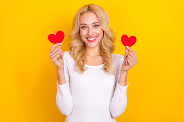 Photo of pretty adorable young woman wear white shirt holding two little hearts isolated yellow color background