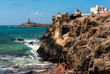 Fototapeta na wymiar Vista de casas antiguas de Puerto de la Cruz que cuelgan en las rocas y el faro de Punta Jandia en el fondo, Fuerteventura, Islas Canarias, España