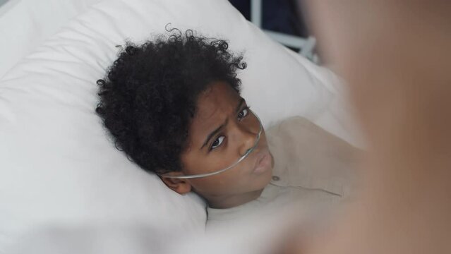 Over shoulder of cropped doctor talking to African American boy with oxygen tube lying in bed in hospital room at daytime