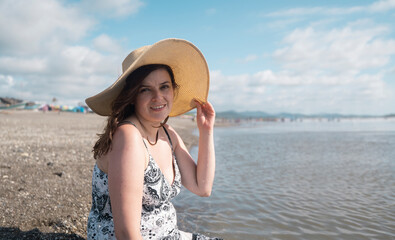 Beautiful happy young Hispanic woman sitting alone at the edge of the beach looking at the camera, wearing a hat and a black and white dress during a sunny morning