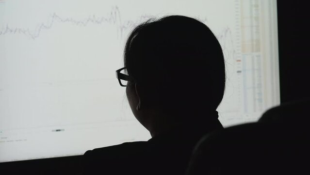 Businesswoman is looking stock data chart to taking note on document and checking data of stock exchange trading on computer while sitting to analysis for play stock in market at office at night.