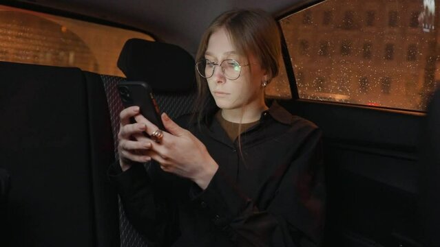 Young woman in glasses with long blond straight hair reads and scroll news on smartphone in a taxi at night. Slow motion 4k footage