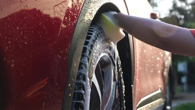 Close-up of a woman's hand washing the alloy wheels of a car with sponge and soap on the car wash. Manual car wash with white soap. Car wash service concept.
