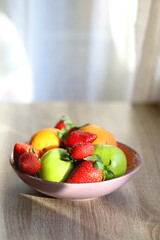 Pink bowl filled with fresh apples, oranges and strawberries on wooden table. Selective focus.