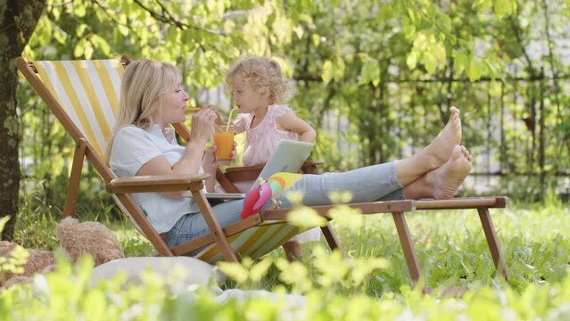 Happy smiling family in green garden, sunny summer day. Mother and her little child girl have fun together playing and drinking orange juice. Mom on break from using laptop spends time with daughter