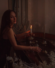 magic medieval castle night. young brunette girl in black silk dress stands in profile near grand piano  and looks apart with candle in hand on a vintage room background. lifestyle concept, free space