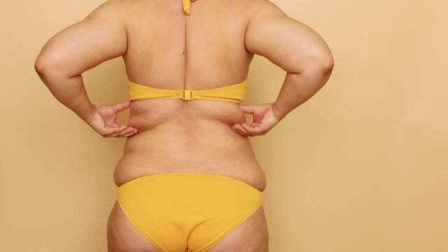 Back view of unrecognizable fat woman wearing yellow bra, bikini, stretching, squeezing excess subcutaneous fat of back, massage on beige background. Body positive, obesity, weight loss, liposuction.
