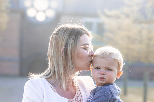 a young happy mother with a small child in her arms, gently kissing his head, stands in the middle of a park in the rays of the rising morning sun. High quality photo