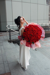 bride celebration. young brunette girl in white slim wedding dress stands casual with huge luxury bouquet of red roses in hands on street stairs gray building background. lifestyle concept, free space