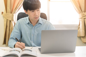 Young Asian college male student writing notes, using laptop for learning online education, business learning, watching online webinar. Businessman is trading, investing on online platform on laptop.