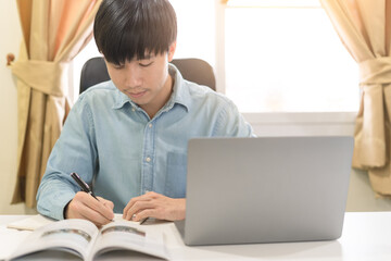 Young Asian college male student writing notes, using laptop for learning online education, business learning, watching online webinar. Businessman is trading, investing on online platform on laptop.