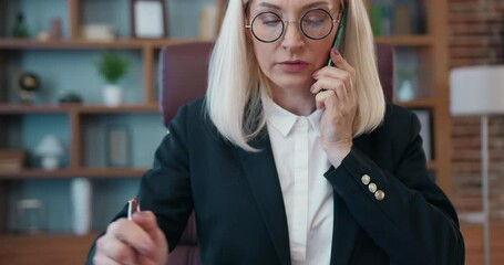 Beautiful smiling older businesswoman is talking on mobile phone and typing on laptop in modern office communicating with business partners. Discuss business issues on phone.