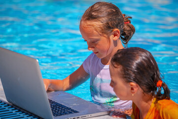 Summer holiday and education concept. Portrait of two young beautiful happy child girls learning remotely with laptop in swimming pool.