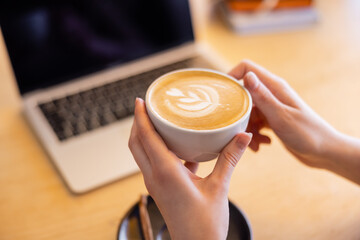 cropped view of young woman holding cup of cappuccino near laptop in cafe.