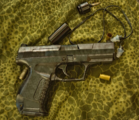Obrazki ze strzelnicy , pistolet , łuski i zatyczki do uszu ( ochronniki słuchu )  leżące na materiale w kamuflażu " żaba - puma " .