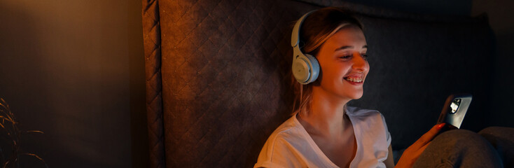 Young woman in headphones using cellphone while sitting on bed