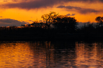 Fototapeta na wymiar Orange purple and violet sunset on river with dark colorful clouds in sky with trees reflection in water 