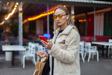 Young fashion woman in yellow sunglasses and earphones using smartphone in evening city