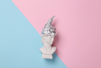 Antique david statue in foil hat on pink blue background. Conspiracy theory