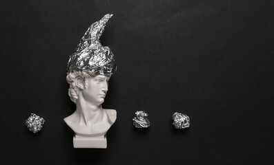 Antique david statue in foil hat on black background. Conspiracy theory - obrazy, fototapety, plakaty