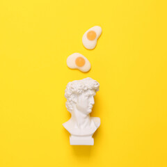 Minimalistic still life. Pop concept. Bust of David with scrambled eggs on a yellow background.