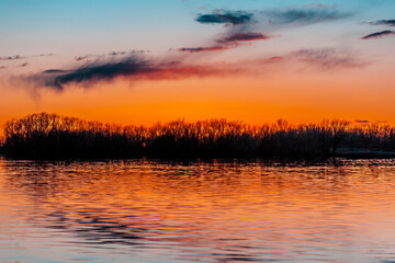Orange purple and violet sunset on river with dark colorful clouds in sky with trees reflection in water 