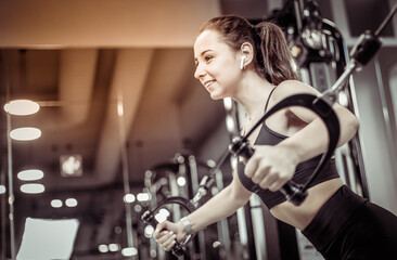 Fototapeta na wymiar Young fit woman trains pectoral misci in a crossover exercise machine. Girl works out in modern gym